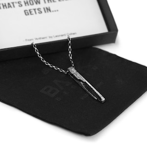 Puede incluir: Un collar de plata con un colgante largo, delgado y texturizado. El colgante tiene forma de barra irregular y dentada. El collar se muestra en una bolsa de terciopelo negro.