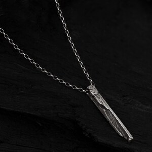 Puede incluir: Un collar de plata con un colgante rectangular largo y texturizado. El colgante tiene un acabado martillado rugoso.