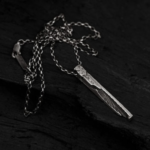 Puede incluir: Un collar de plata con un colgante rectangular largo y texturizado. El colgante tiene un acabado rugoso y martillado.