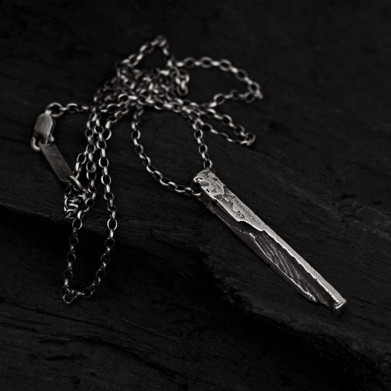 Puede incluir: Un collar de plata con un colgante rectangular largo y texturizado. El colgante tiene un acabado rugoso y martillado.