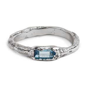 May include: A silver ring with a textured band and a rectangular, light blue gemstone. The ring's design features a rough, organic texture, giving it a unique, handcrafted appearance. The gemstone is set in a bezel setting.