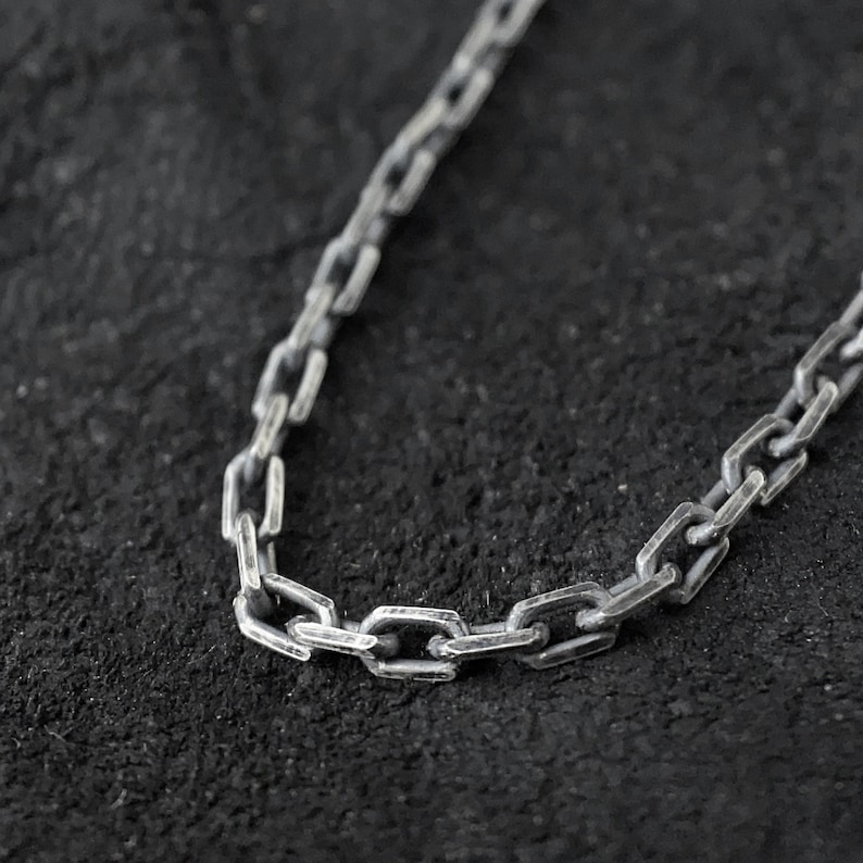 May include: A silver chain necklace with a box chain design. The chain is made of small, rectangular links.