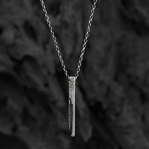Puede incluir: Un collar de cadena de plata con un colgante de barra vertical texturizado. El colgante tiene un aspecto &aacute;spero y org&aacute;nico.