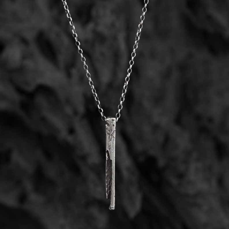Puede incluir: Un collar de cadena de plata con un colgante de barra vertical texturizado. El colgante tiene un aspecto &aacute;spero y org&aacute;nico.