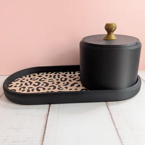 Könnte beinhalten: Ein ovales schwarzes Tablett mit einem beigefarbenen und schwarzen Leopardenmuster als Boden. Ein zylindrischer schwarzer Behälter mit passendem Deckel und einem kleinen goldfarbenen Knopf steht auf der rechten Seite des Tabletts.
