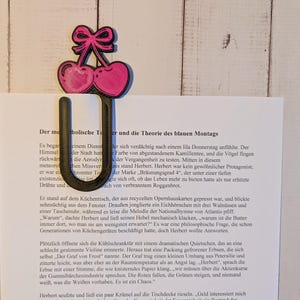 Könnte beinhalten: Eine schwarze Büroklammer aus Metall mit einem Design aus rosa Kirschen. Die Kirschen haben eine rosa Schleife oben. Die Klammer hält ein weißes Blatt Papier mit deutschem Text.