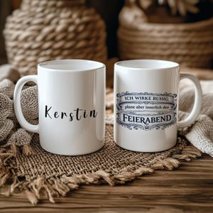 Könnte beinhalten: Zwei weiße Keramikbecher. Ein Becher hat den Namen "Kerstin" in schwarzer Schrift. Der andere Becher hat den Text "ICH WIRKE RUHIG, plane aber innerlich den FEIERABEND" in blauer Schrift in einem dekorativen Rahmen. Beide Becher haben einen gebogenen Griff.