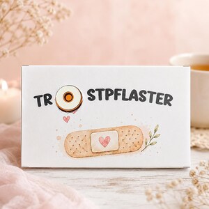 Peut inclure: Une boîte rectangulaire blanche portant le texte TROSTPFLASTER en lettres noires. La lettre O est remplacée par une illustration d'œuf au plat. En dessous, un pansement adhésif beige affiche un petit cœur rose et un brin de feuilles vertes.