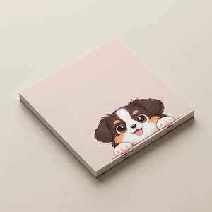 Könnte beinhalten: Ein quadratischer, hellrosa Notizblock mit einer Cartoon-Welpen-Illustration. Der Welpe hat braunes und weißes Fell, große braune Augen und eine rosa Zunge. Der Notizblock ist ein Stapel Papier, ideal zum Notieren.