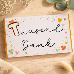 Könnte beinhalten: Eine rechteckige weiße Geschenkbox mit dem Text Tausend Dank in schwarzer Schreibschrift. Die Box ist mit kleinen bunten Herzen und Sternen in Orange, Rosa und Grün verziert. Ein großer ausgeschnittener Buchstabe T befindet sich links.