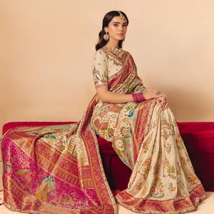 May include: An ivory-colored sari with a vibrant pink border and intricate patterns. The sari features detailed illustrations of figures, plants, and animals. The design includes a matching blouse and is adorned with gold and red accents.