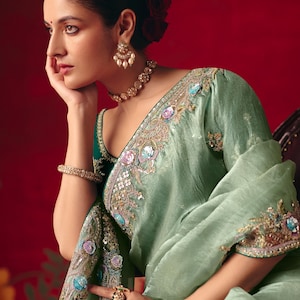 May include: A light sage green saree featuring intricate embroidery with sequins and floral motifs in shades of blue, pink, and gold. The garment is paired with a dark green blouse, a gold-toned choker necklace, matching drop earrings, and a bracelet.