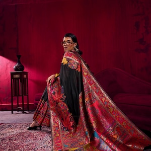 May include: A black shawl with detailed floral and paisley patterns in red, yellow, and blue. The shawl is draped over the shoulders, displaying the detailed embroidery and rich colours. The background is a deep red.
