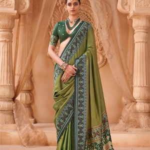 Op de afbeelding: Een olijfgroene sari met een donkergroene rand voorzien van complexe geometrische patronen en olifantenmotieven. De bijpassende blouse heeft korte mouwen en een diepe halslijn. Het geheel is afgewerkt met een meerlaagse kralenketting en armbanden.