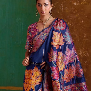 May include: A navy blue saree featuring a vibrant, multi-colored leaf pattern in shades of orange, pink, and yellow. The garment includes a matching blouse with a dense, intricate floral design and a wide, ornate border along the edges.
