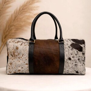 Pu&ograve; includere: Una borsa da viaggio rettangolare con un motivo a pelle di mucca. La borsa presenta un pannello centrale marrone, affiancato da pannelli di pelle maculata bianca e marrone. Manici e finiture in pelle nera completano il design.