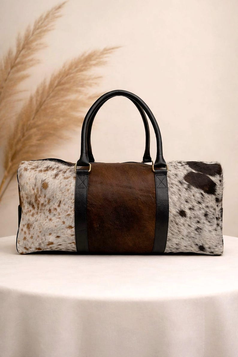 Pu&ograve; includere: Una borsa da viaggio rettangolare con un motivo a pelle di mucca. La borsa presenta un pannello centrale marrone, affiancato da pannelli di pelle maculata bianca e marrone. Manici e finiture in pelle nera completano il design.