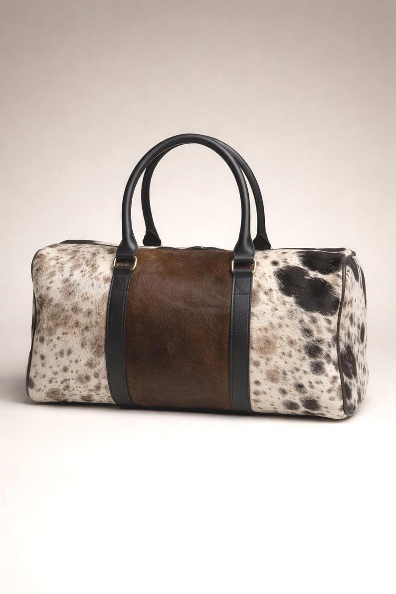 May include: A rectangular duffel bag with a cowhide pattern in shades of brown, black, and white. The centre panel is solid brown, flanked by black leather straps. It has two black leather handles.