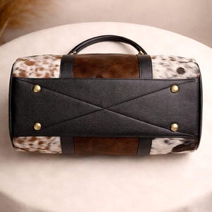 Pu&ograve; includere: Borsa da viaggio rettangolare con motivo a pelle di mucca marrone e bianca. La borsa presenta una base e dettagli in pelle nera, con un manico superiore e rivetti color oro. Progettata per i viaggi.