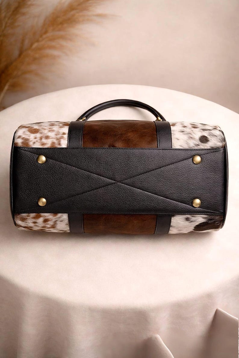 Pu&ograve; includere: Borsa da viaggio rettangolare con motivo a pelle di mucca marrone e bianca. La borsa presenta una base e dettagli in pelle nera, con un manico superiore e rivetti color oro. Progettata per i viaggi.