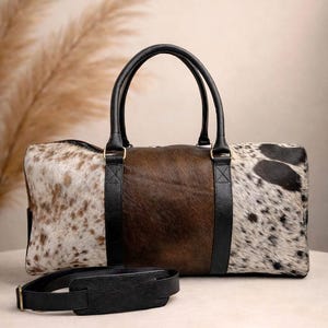 Pu&ograve; includere: Una borsa da viaggio in pelle di mucca con manici e dettagli in pelle nera. La borsa presenta un pannello centrale marrone, affiancato da pannelli in pelle maculata bianca e marrone. Una tracolla nera &egrave; davanti alla borsa.