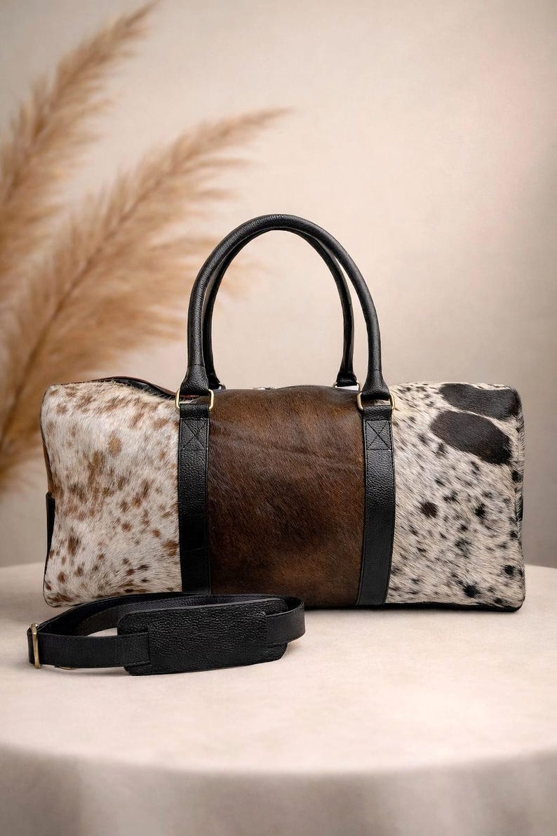 Pu&ograve; includere: Una borsa da viaggio in pelle di mucca con manici e dettagli in pelle nera. La borsa presenta un pannello centrale marrone, affiancato da pannelli in pelle maculata bianca e marrone. Una tracolla nera &egrave; davanti alla borsa.