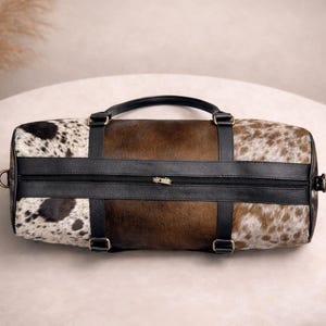 Pu&ograve; includere: Una borsa da viaggio cilindrica con un motivo a pelle di mucca. La borsa presenta una combinazione di sezioni marroni, nere e bianche macchiate. Ha un manico e cinghie in pelle nera e una chiusura con cerniera. La borsa &egrave; progettata per viaggiare o trasportare oggetti.