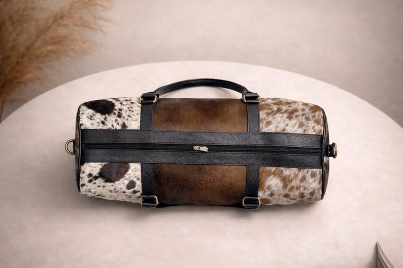 Pu&ograve; includere: Una borsa da viaggio cilindrica con un motivo a pelle di mucca. La borsa presenta una combinazione di sezioni marroni, nere e bianche macchiate. Ha un manico e cinghie in pelle nera e una chiusura con cerniera. La borsa &egrave; progettata per viaggiare o trasportare oggetti.