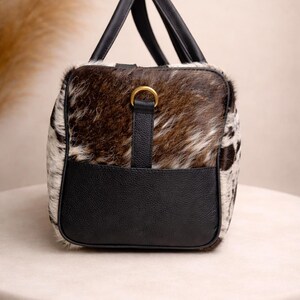 Pu&ograve; includere: Una borsa da viaggio rettangolare con motivo a pelle di mucca e dettagli in pelle nera. La borsa presenta una combinazione di pelliccia marrone, bianca e nera, con manici in pelle nera e una base in pelle nera. &Egrave; presente un dettaglio ad anello color oro.