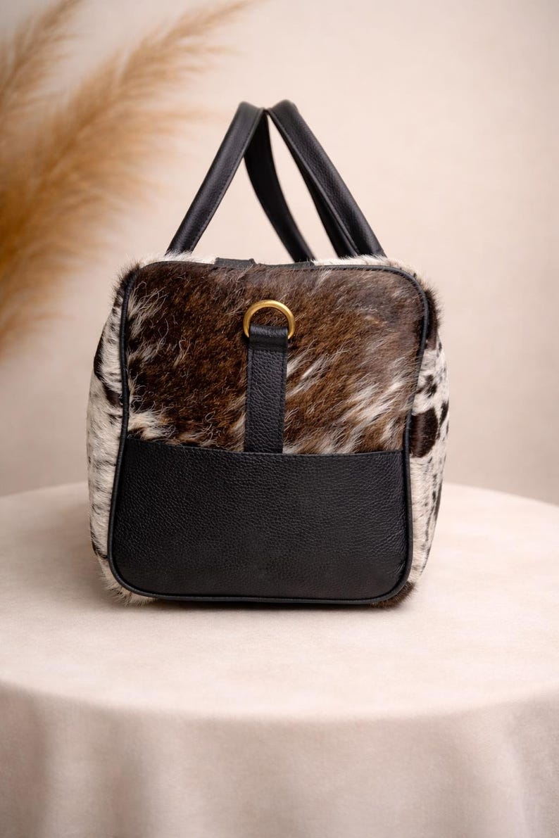 Pu&ograve; includere: Una borsa da viaggio rettangolare con motivo a pelle di mucca e dettagli in pelle nera. La borsa presenta una combinazione di pelliccia marrone, bianca e nera, con manici in pelle nera e una base in pelle nera. &Egrave; presente un dettaglio ad anello color oro.
