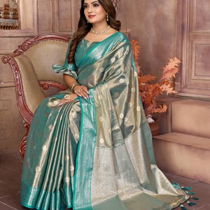 May include: A shimmering, teal and gold saree with intricate detailing. The fabric has a metallic sheen, with a teal border and tassels. The saree is draped over a chair, showcasing its elegant design and texture.