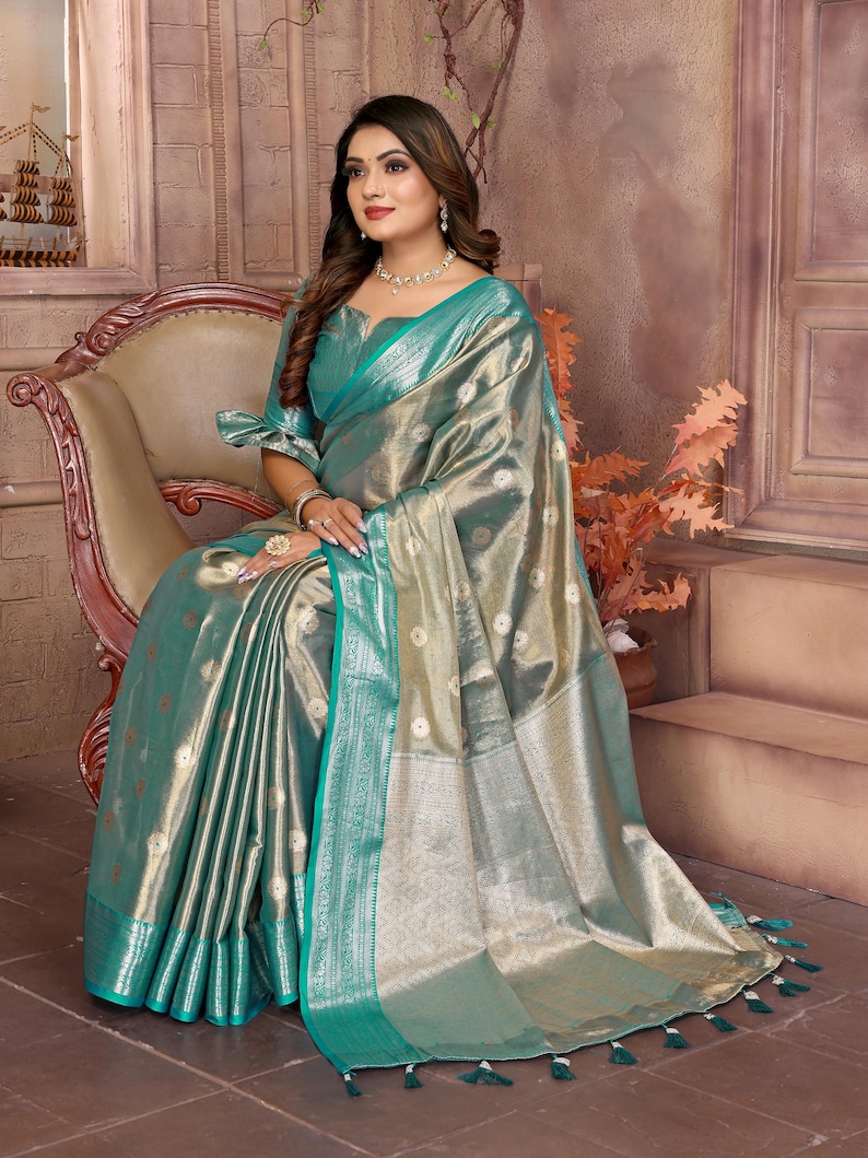 May include: A shimmering, teal and gold saree with intricate detailing. The fabric has a metallic sheen, with a teal border and tassels. The saree is draped over a chair, showcasing its elegant design and texture.
