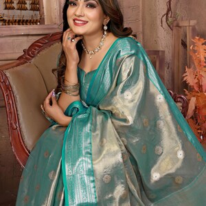 May include: A woman wearing a teal and gold sari, seated in a wooden chair. The sari features a shimmering fabric with floral patterns and a teal border. She is adorned with gold jewelry, including a necklace, earrings, and bracelets. The background includes a decorative wooden ship.