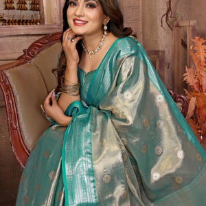 May include: A woman wearing a teal and gold sari, seated in a wooden chair. The sari features a shimmering fabric with floral patterns and a teal border. She is adorned with gold jewelry, including a necklace, earrings, and bracelets. The background includes a decorative wooden ship.