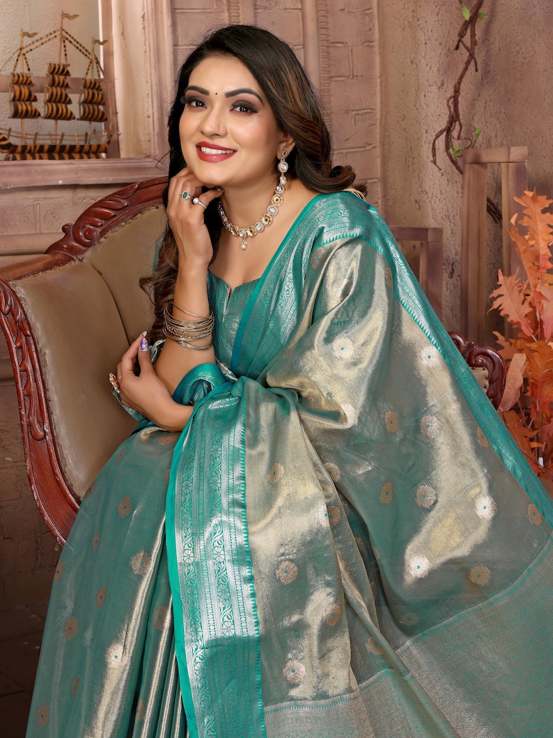 May include: A woman wearing a teal and gold sari, seated in a wooden chair. The sari features a shimmering fabric with floral patterns and a teal border. She is adorned with gold jewelry, including a necklace, earrings, and bracelets. The background includes a decorative wooden ship.