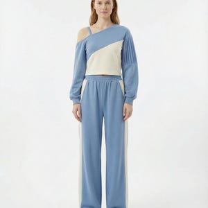 May include: A two-piece outfit featuring a light blue and off-white top with an asymmetrical shoulder design and pleated sleeve detail. The matching light blue pants have a wide leg and off-white side stripes. The outfit is made of a soft material.