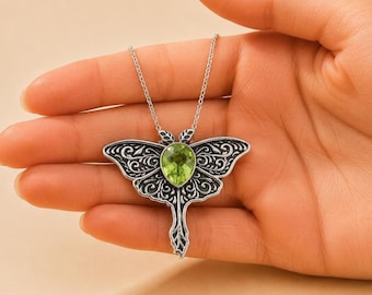 May include: A silver-toned metal pendant shaped like a moth with intricate filigree patterns and a central pear-shaped lime green gemstone. The pendant hangs from a thin silver-toned chain, showcasing detailed wing textures and a pointed tail.