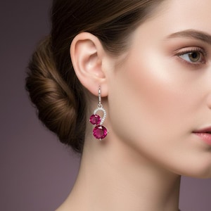 Può includere: Eleganti orecchini in argento con due grandi pietre preziose rotonde color rosso rubino. Gli orecchini presentano un design curvo con un piccolo accento di pietre preziose rotonde e chiare. Il gioiello è su uno sfondo sfocato.