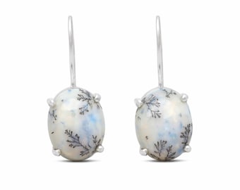 May include: A pair of drop earrings featuring oval-shaped dendritic agate stones set in silver prongs. The stones are white with translucent blue patches and dark, fern-like inclusions. Each earring hangs from a simple silver hook wire.