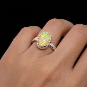 Könnte beinhalten: Ein silberner Ring mit einem ovalen Opal-Edelstein. Der Opal zeigt eine Mischung aus Gelb, Grün und Orange. Der Ring hat ein schlichtes, poliertes Band und eine Lünettenfassung, die den Opal sicher hält.