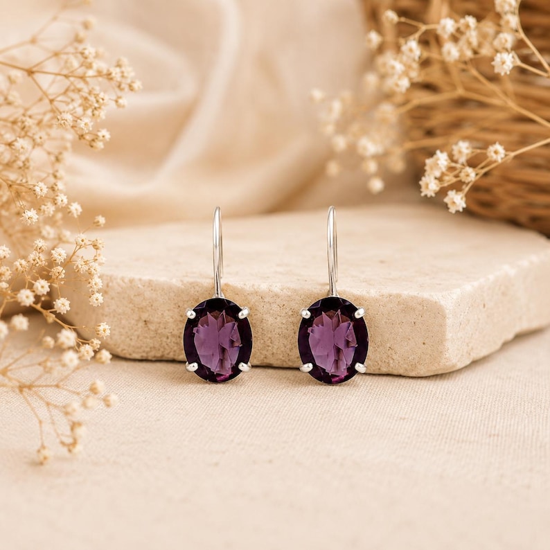 May include: A pair of silver drop earrings featuring oval-cut deep purple amethyst gemstones. Each stone is secured in a four-prong setting attached to a simple silver hook wire, showcasing a faceted surface that reflects light clearly.