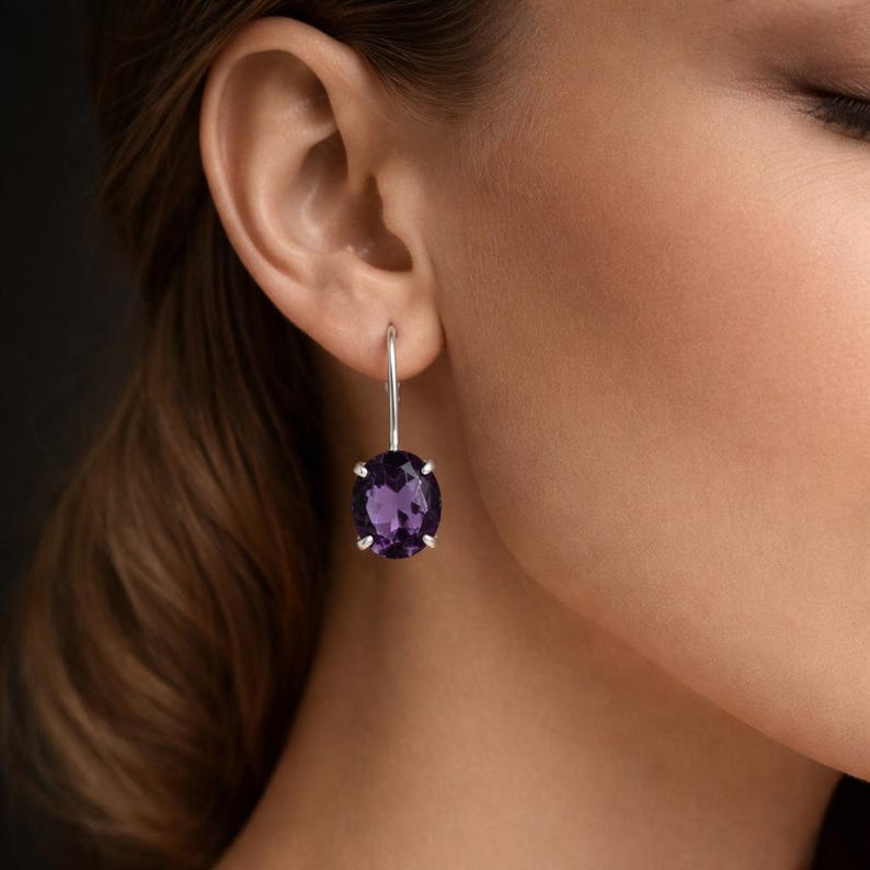 May include: A silver drop earring featuring a single oval-cut deep purple amethyst gemstone held by four prongs. The earring hangs from a simple silver hook mechanism, showcasing the faceted stone's clarity and rich violet color against the skin.
