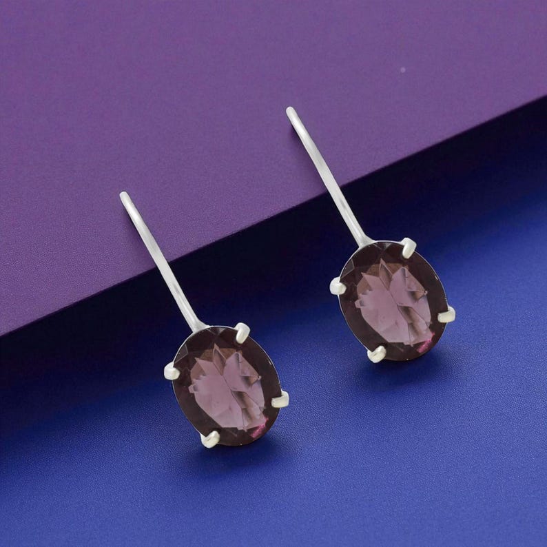 May include: A pair of silver drop earrings featuring oval-shaped, translucent purple gemstones. Each stone is secured by four silver prongs and attached to a thin, straight silver post that extends upward from the top of the setting.
