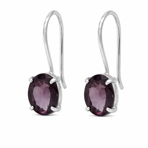 May include: A pair of drop earrings featuring oval-cut, deep purple gemstones set in polished silver prongs. Each earring is suspended from a simple, curved silver hook wire, showcasing a faceted surface that reflects light across the dark purple stone.