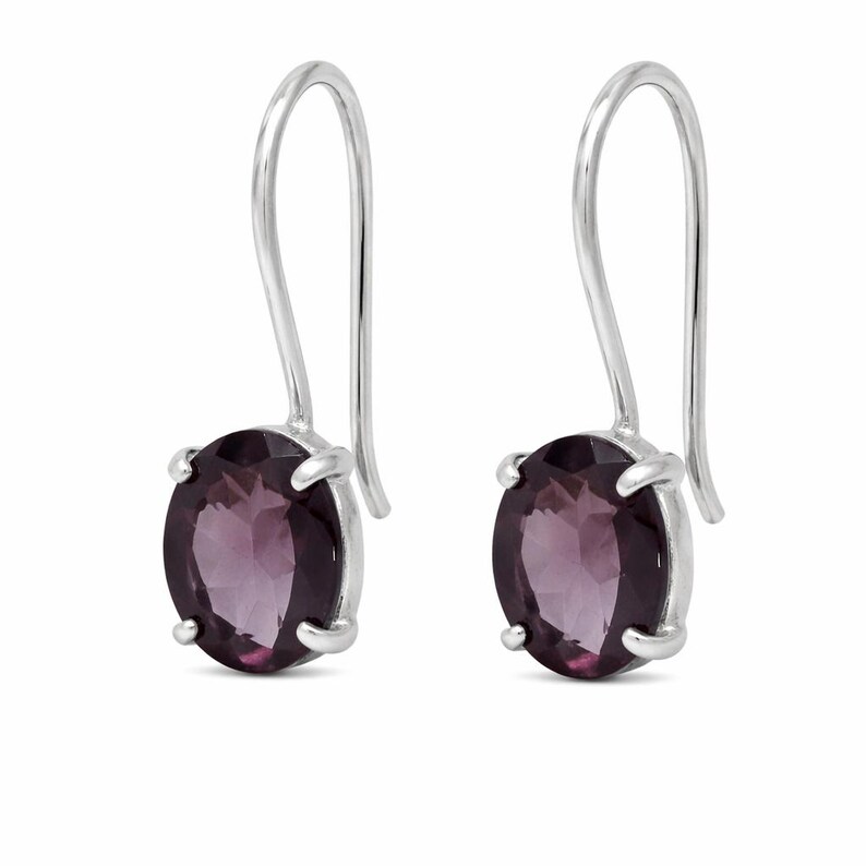 May include: A pair of drop earrings featuring oval-cut, deep purple gemstones set in polished silver prongs. Each earring is suspended from a simple, curved silver hook wire, showcasing a faceted surface that reflects light across the dark purple stone.