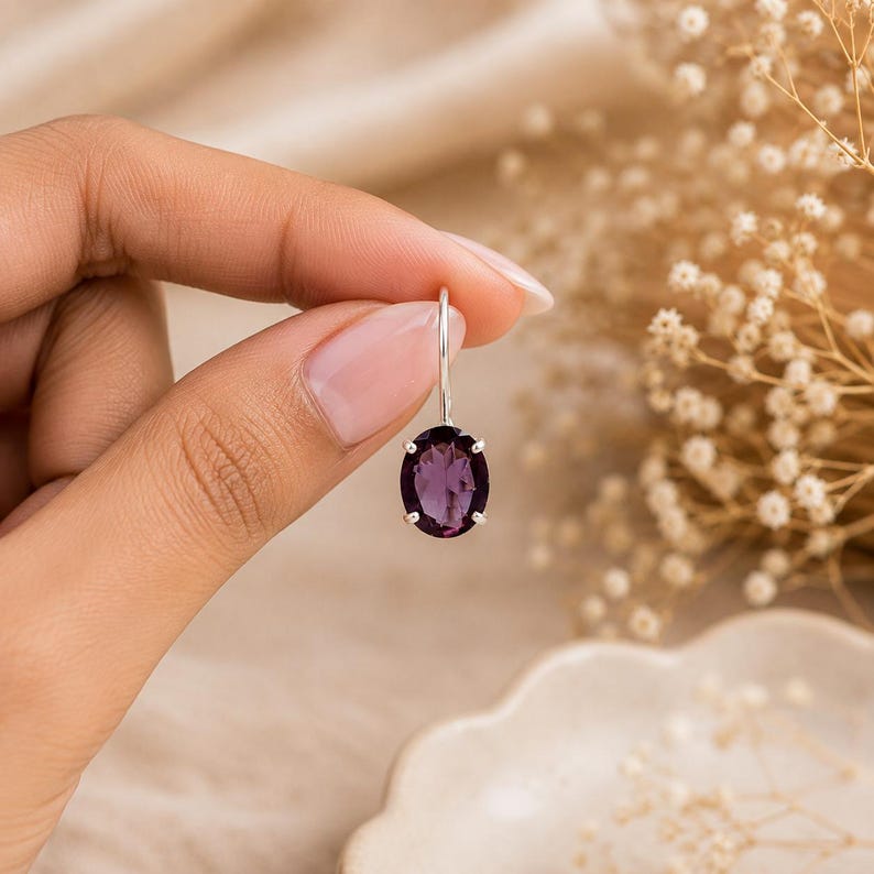 May include: A single oval-cut purple amethyst gemstone earring set in a silver prong setting. The earring features a simple silver hook wire for attachment, showcasing the deep violet color and faceted surface of the stone in a clear, bright light.