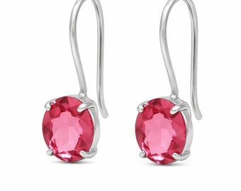 May include: A pair of silver drop earrings featuring oval-cut, bright pink gemstones. The gemstones are set in a silver bezel and suspended from a curved silver hook. The earrings are displayed against a white background.