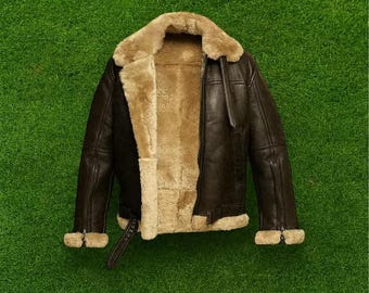May include: A brown leather jacket with a tan shearling lining and collar. The jacket has a zipper closure, a buckled belt at the waist, and shearling trim at the cuffs. The jacket is displayed on a green background.