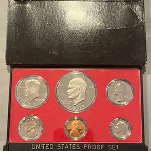 May include: A 1973 United States Proof Set in a black case. The set includes six coins of various sizes, including a silver dollar, half dollar, quarter, dime, nickel, and penny. The coins are displayed on a red background.