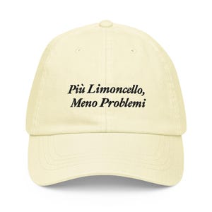 May include: A cream-colored baseball cap with a curved brim. The cap features the text "Più Limoncello, Meno Problemi" in black, centered on the front. The hat has a classic six-panel design with a top button.